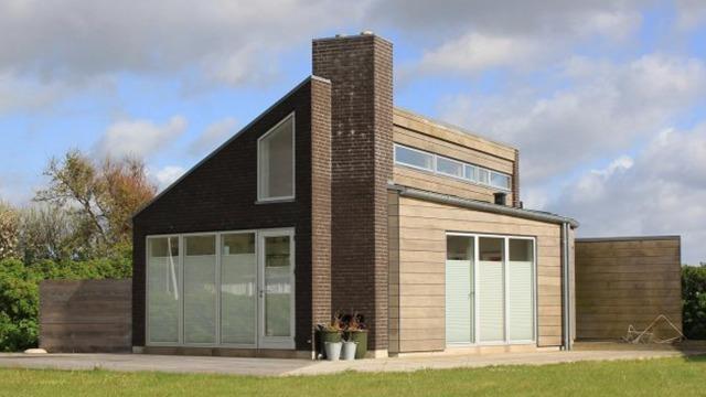 Moderne hus med træfacade og store vinduer, beliggende i et grønt område under en skyet himmel.