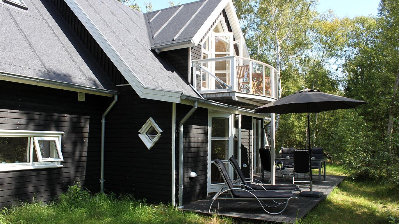 Sort sommerhus med terrasse og parasol omgivet af grønne træer.