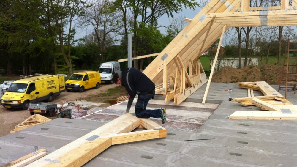 En håndværker arbejder på taget af et hus, mens byggepladsen er fyldt med lastbiler og træmaterialer.