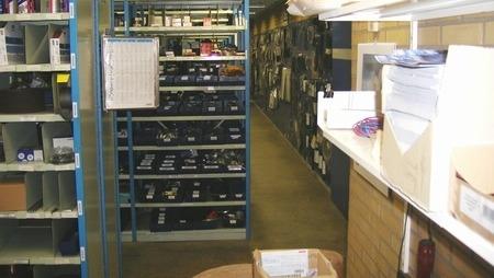 Lagerlokale med reoler fyldt med kasser og materialer samt en arbejdsstation med papirer og kontorartikler.