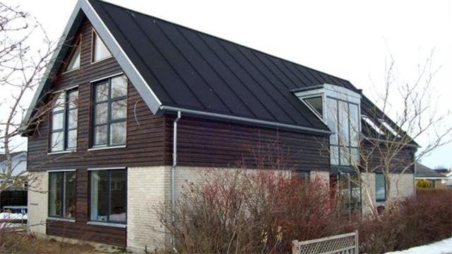 Moderne hus med sort tag og træfacade, omgivet af buske og træer i en rolig boligområde.