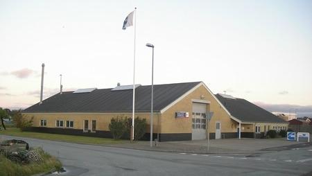 En gul bygning med sort sokkel og flagstang, beliggende ved en vej i et roligt område.