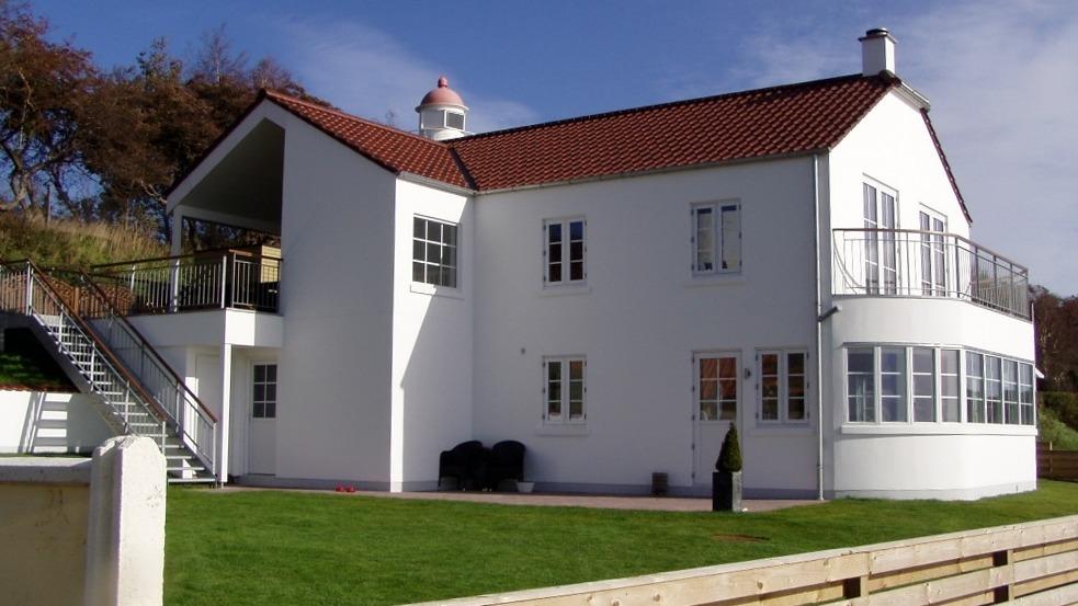 Et moderne hvidt hus med rød tag, balkon og stor græsplæne under en blå himmel.