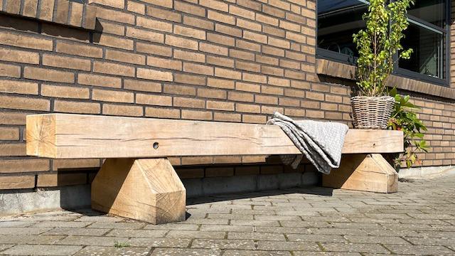 Bænk i træ med en plaid og en plante i en kurv, placeret ved en mur af mursten.