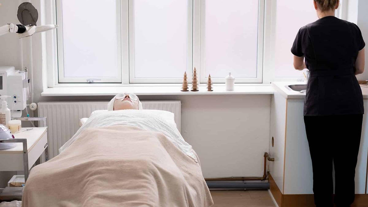 En behandler står ved vasken, mens en patient ligger på en seng i et lyst behandlingsrum.
