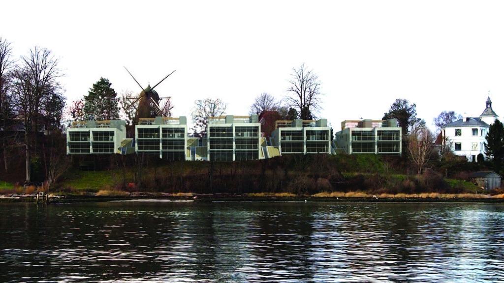 Moderne boliger ved vandet med en vindmølle i baggrunden og træer langs kystlinjen.