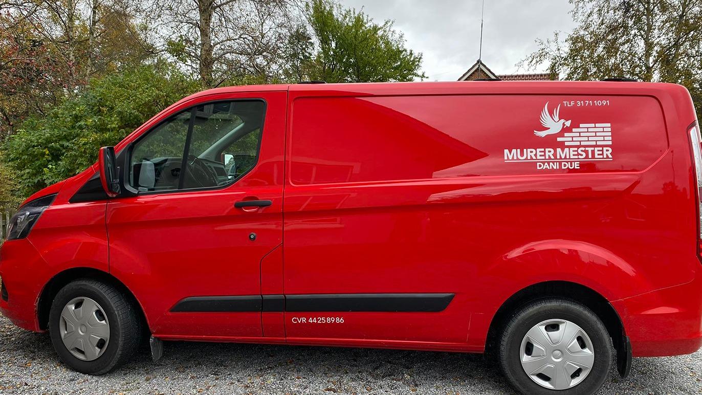 Rød varevogn med teksten "Murer Mester" på siden, parkeret på grusvej omgivet af træer.