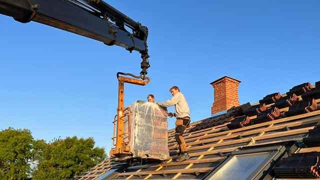 To personer arbejder på et tag med en kran, der løfter materialer under en klar blå himmel.