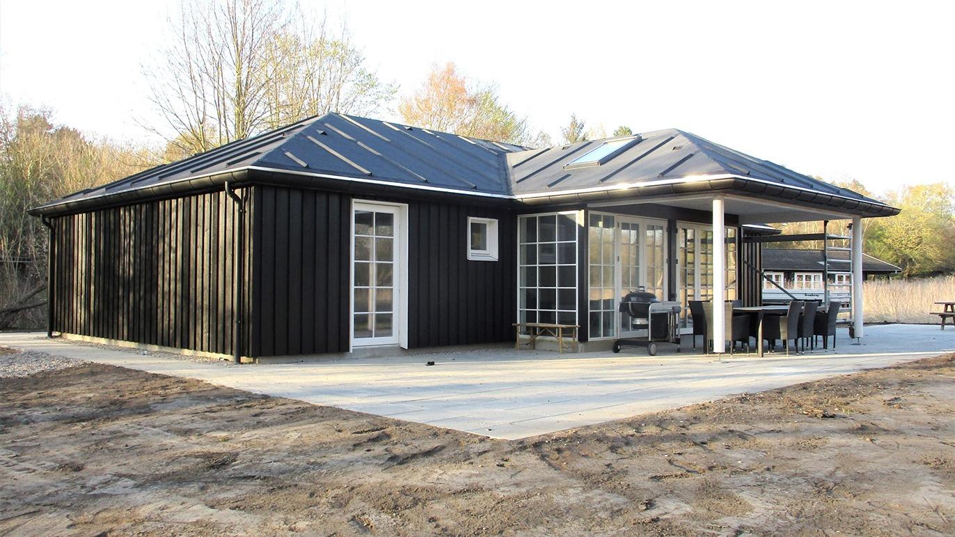 Moderne sort hus med terrasse og glasvæg, beliggende i et naturskønt område.