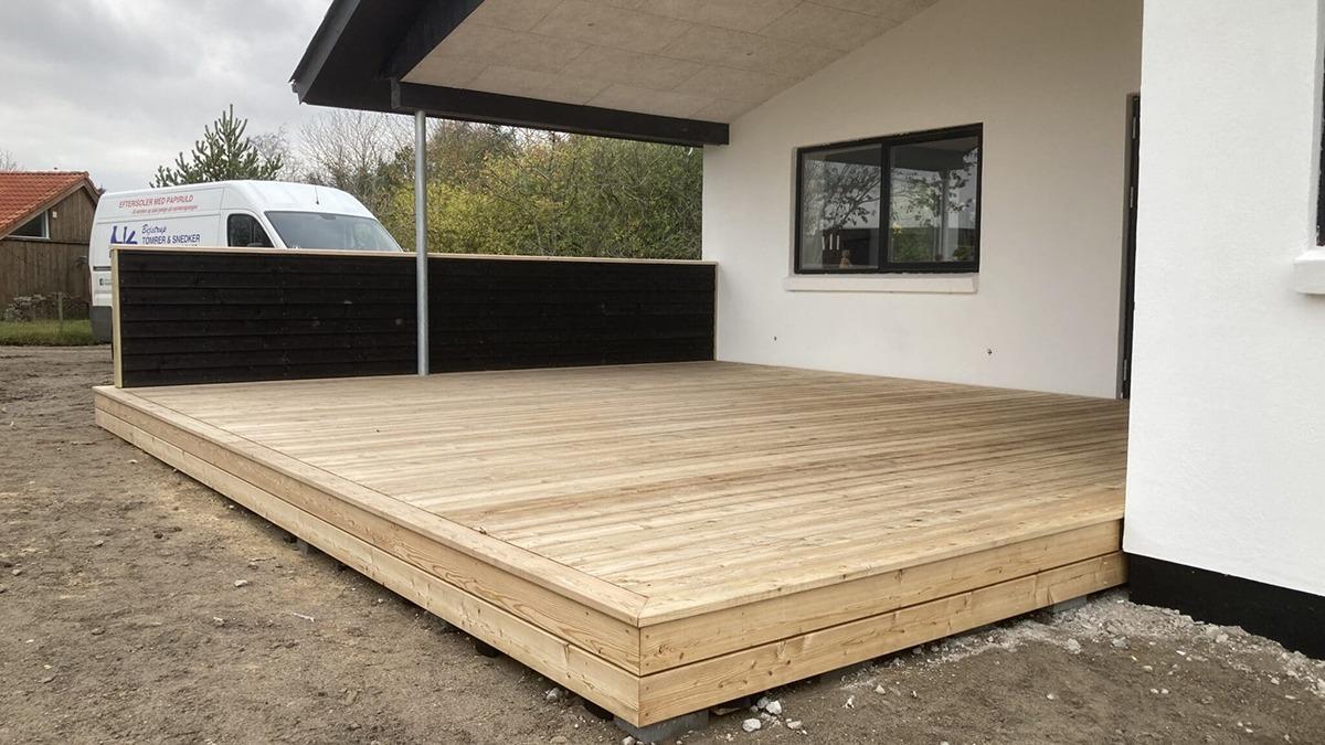 En ny træterrasse foran et hus med en sort væg og en hvid facade, delvist dækket af en grå himmel.