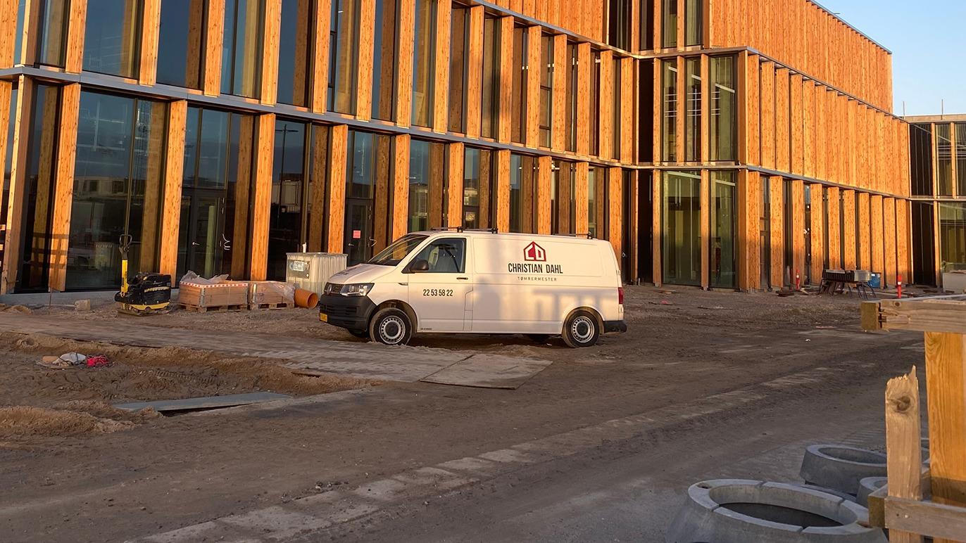 En hvid varevogn parkeret foran en moderne bygning med træfacade og byggeplads i forgrunden.