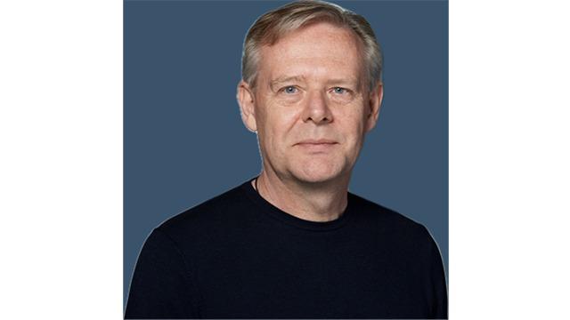 En mand med kort, lyst hår iført en sort trøje står foran en blå baggrund.