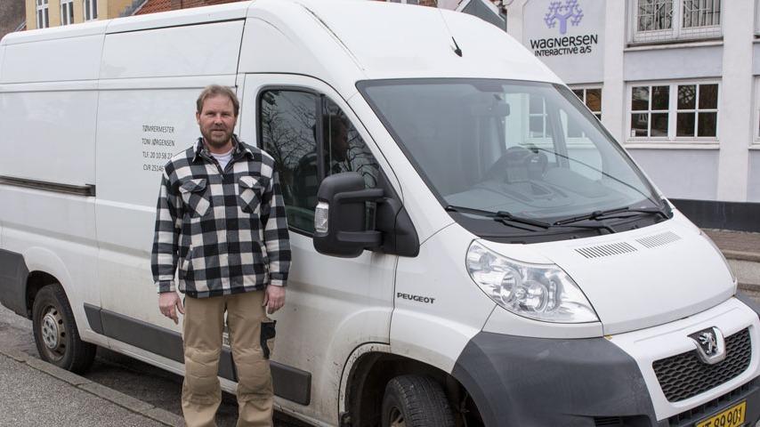 En mand står ved siden af en hvid varevogn på en gade, iført en ternet skjorte og beige bukser.