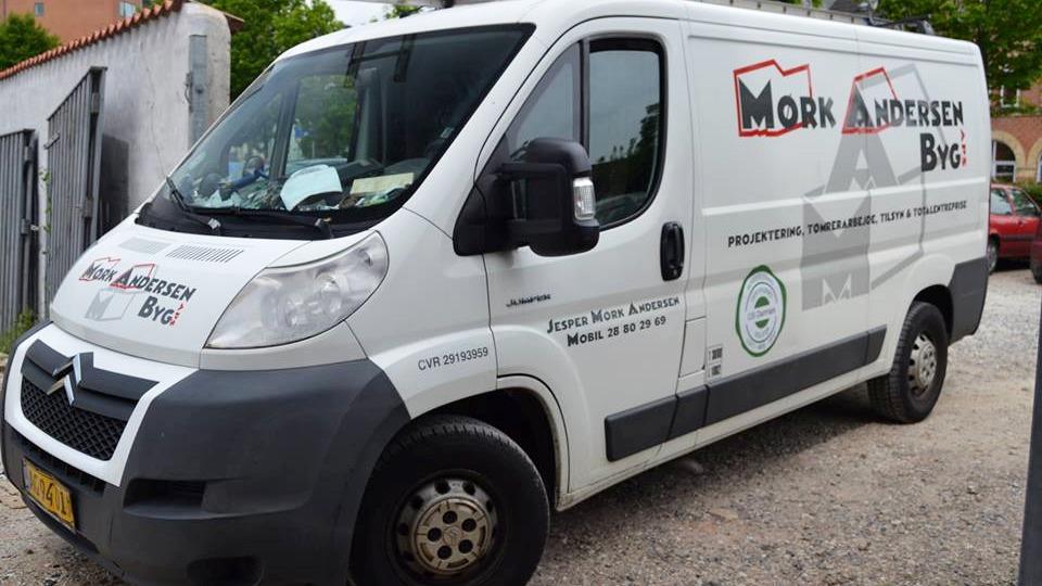 En hvid varevogn med teksten "Mork Andersen Brg" og logoer på siden, parkeret på en grusvej.