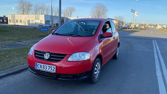 En rød Volkswagen bil parkeret på en vej med bygninger i baggrunden.