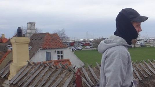 Mand iført grå hættetrøje og sort kasket, går på tag med udsigt over bygninger og landskab.