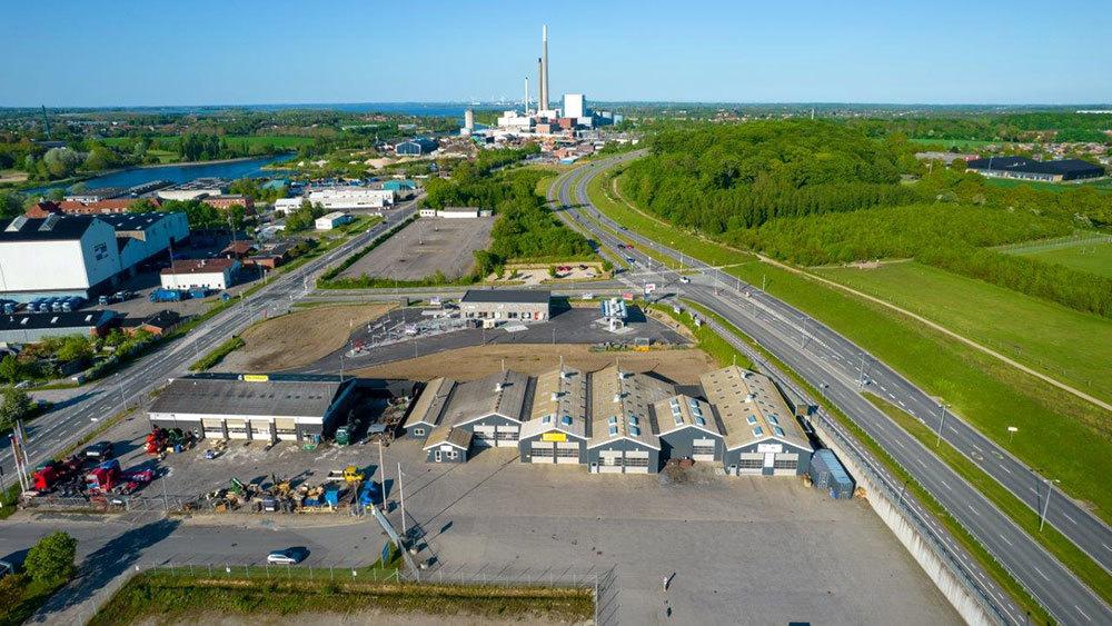 Luftfoto af et industriområde med bygninger, veje og en kraftværk i baggrunden. Grønne områder og vand ses også.