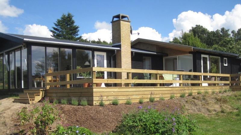 Et moderne hus med træterrasse og skorsten, omgivet af grønne områder og blomster.