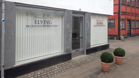 Begravelsesforretning Elving med store vinduer og planter foran i en bygade.