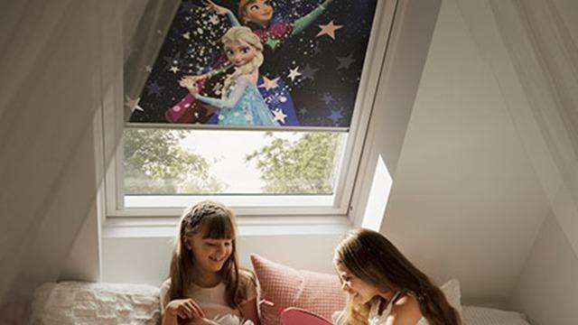 To piger sidder på en seng og leger med en dukke, mens en Frozen-gardine hænger i vinduet.