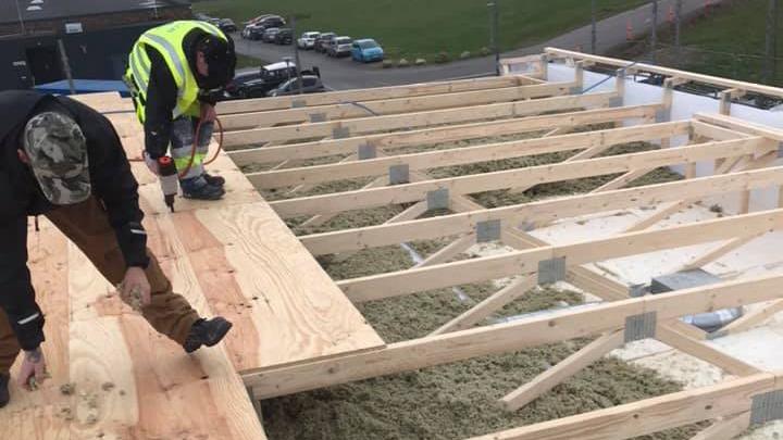 To personer arbejder på et tag med træbjælker og isolering, iført sikkerhedsveste og arbejdstøj.