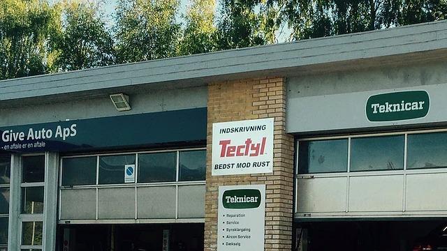 Auto værksted med skilte for Give Auto ApS og Teknicar, beliggende i et område med grønne træer.