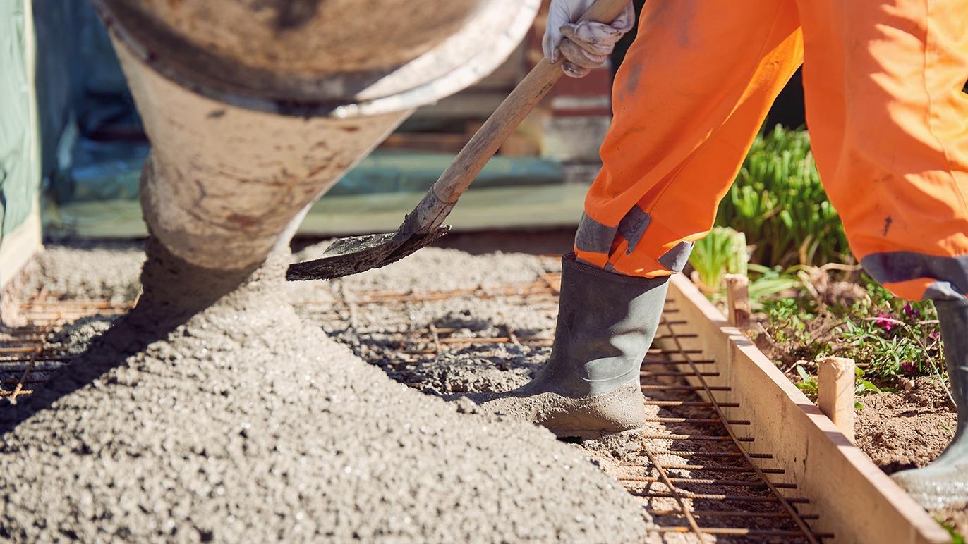 En arbejder hælder beton fra en rørledning og bruger en skovl til at fordele det på byggepladsen.