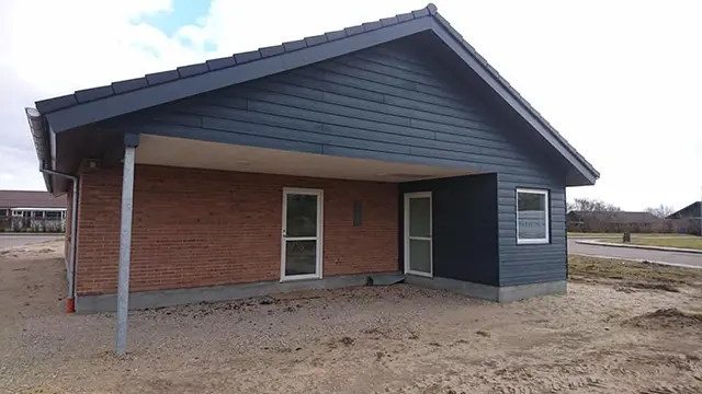 En moderne enetages hus med mørk facade og grå tag, beliggende på en grusvej.