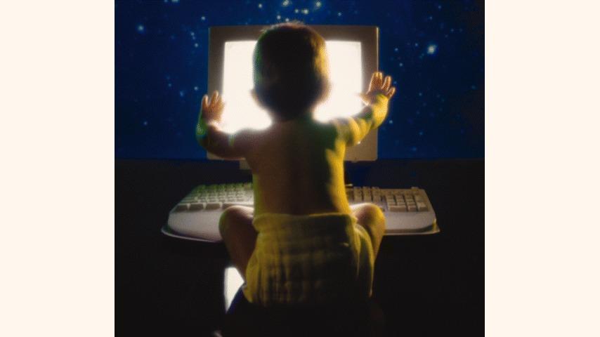 Et spædbarn sidder på gulvet og kigger på en computer med lysende skærm i et mørkt rum.