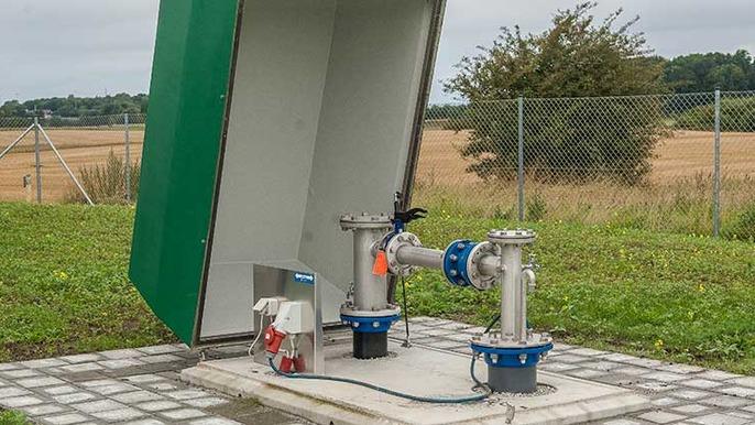 Vandpumpeanlæg med metalrør og grøn skur, beliggende i et åbent landskab med marker i baggrunden.