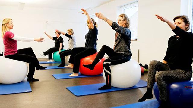 Gruppe af kvinder, der deltager i en pilatesklasse på bold, med fokus på stræk og balanceøvelser.