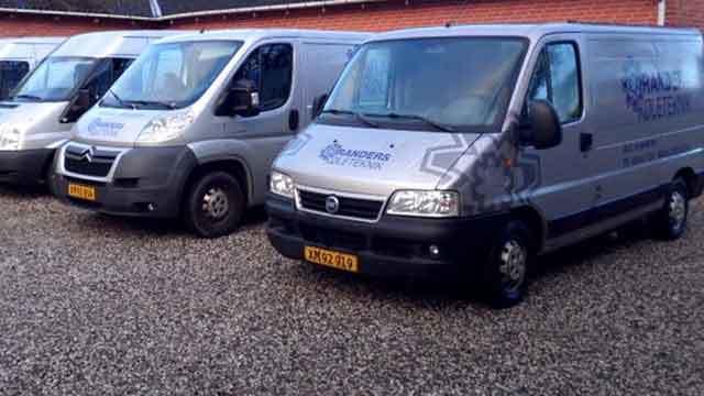 Flere grå varevogne parkeret på en grusplads, med tydelige logoer på siden.