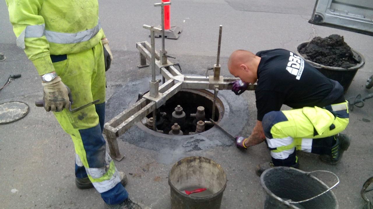 To arbejdere i sikkerhedstøj arbejder ved en brønd, mens de bruger værktøj og en spand.
