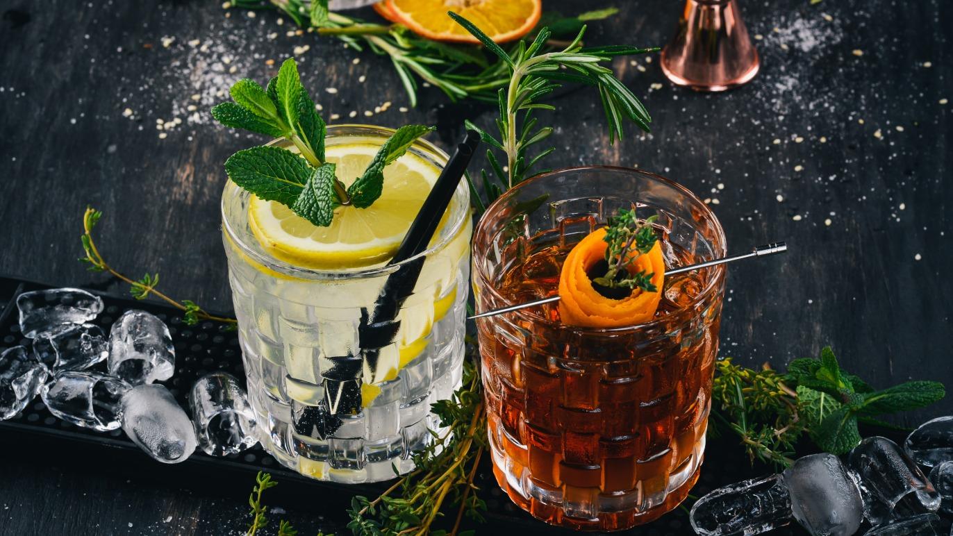 To glas med cocktails, en med mynte og citron, den anden med appelsinskal og urter, på en mørk overflade.