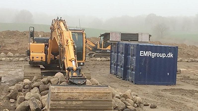 Gul gravemaskine arbejder på en byggeplads med en container og maskiner i baggrunden i tåget vejr.