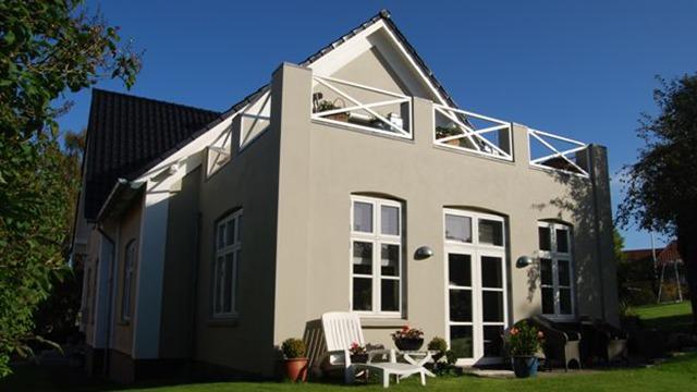Moderne hus med terrasse og store vinduer, beliggende i en grøn have under en klar blå himmel.
