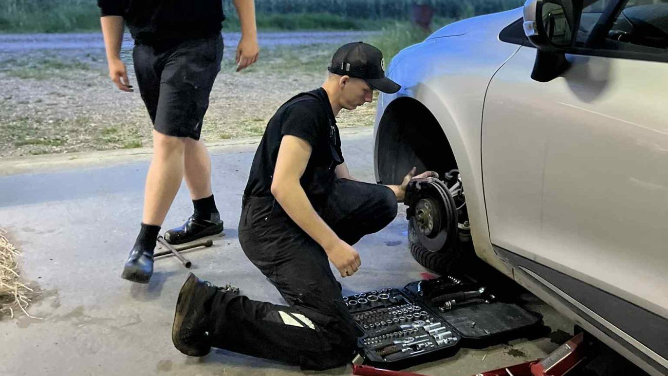 To personer arbejder på at skifte dæk på en bil i en garage, med værktøj spredt på gulvet.
