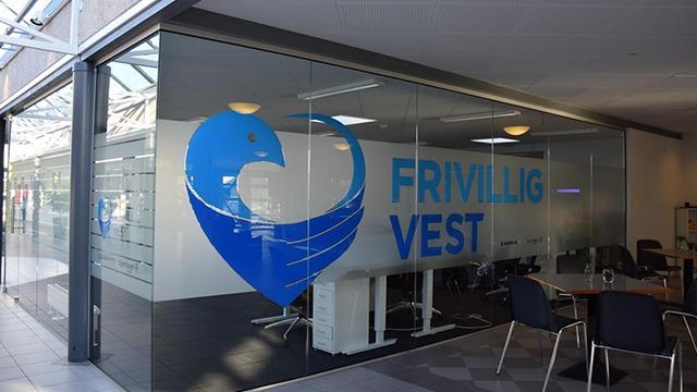 Kontor med glasvæg og logo for Frivillig Vest i blå farver, møbleret med borde og stole.