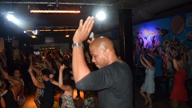 Mand danser med hænderne oppe i en livlig natklub med mange mennesker omkring.