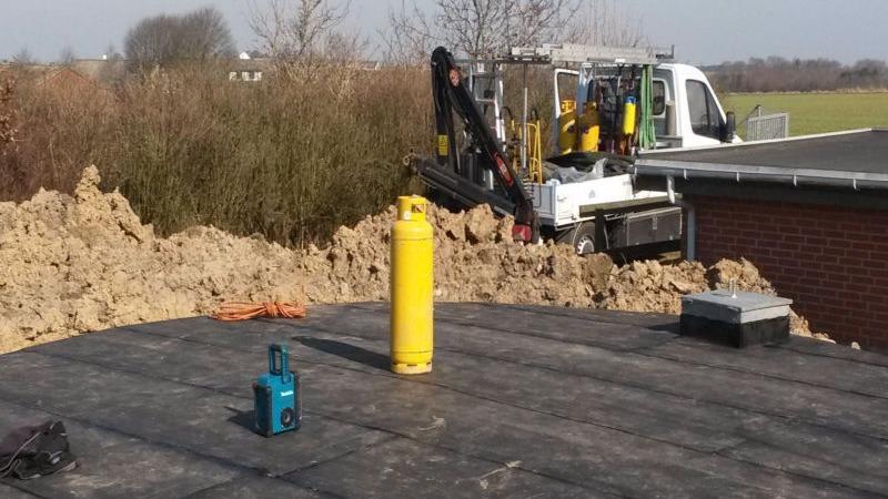 Arbejdsplads med en lastbil, en gul gasflaske og en blå maskine på taget af en bygning, omgivet af gravet jord.