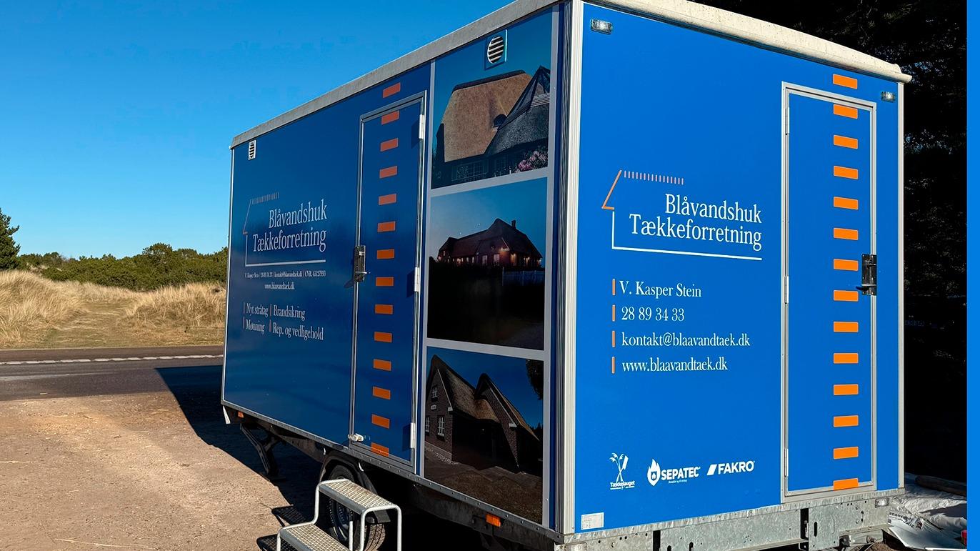 Blå trailer med reklame for Blåvandshuk Tømrerforretning, parkeret ved en vej i naturen.