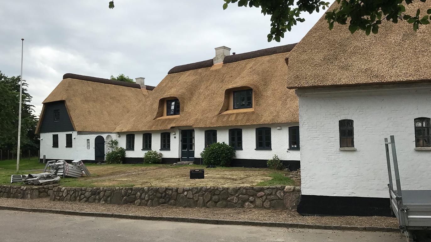 Et smukt hus med stråtag og hvid facade, omgivet af en græsplæne og stenmur.