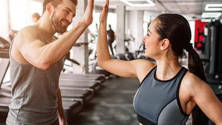 To personer i fitnesscenter, der giver high five efter træning, med træningsudstyr i baggrunden.