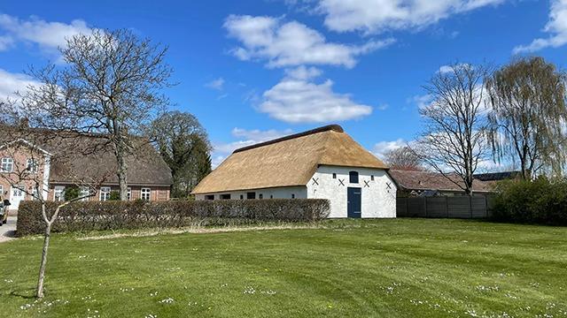 Et hvidt hus med stråtag i en grøn have under en blå himmel med skyer.