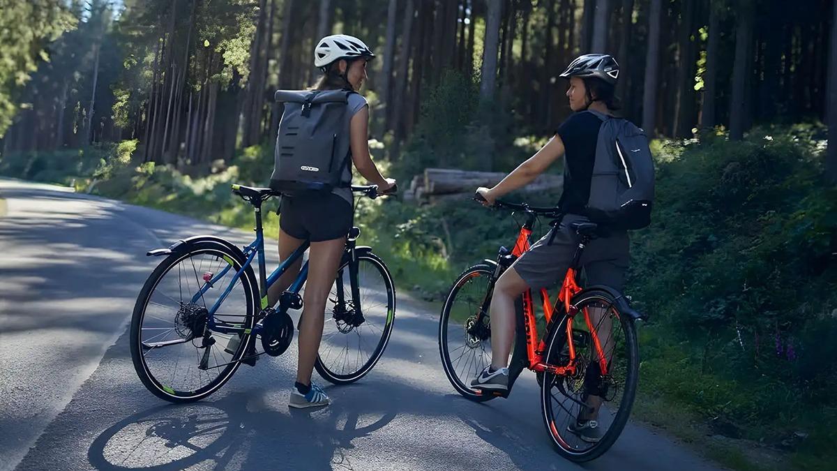 To personer på cykler i skoven, iført hjelme og rygsække, der snakker på en grusvej.