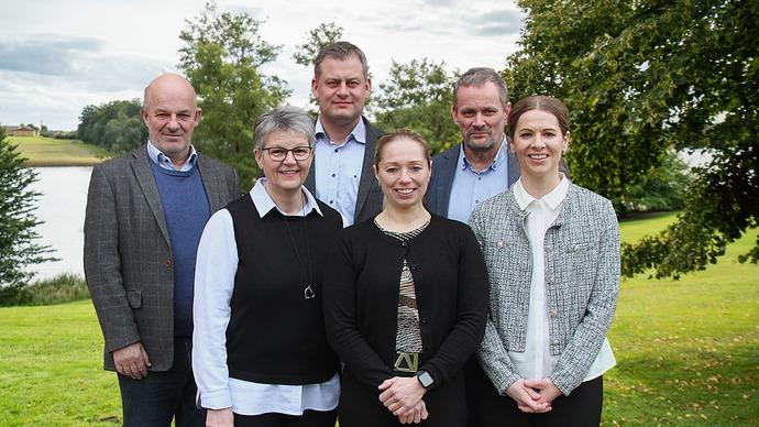 En gruppe på seks personer står sammen på en græsplæne ved en sø, omgivet af træer og en skyet himmel.