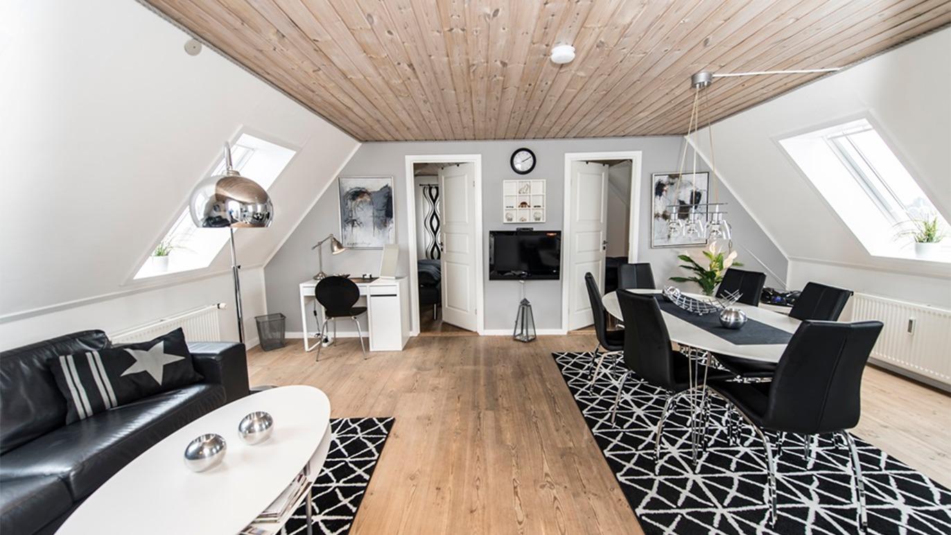Moderne stue med sort-hvide møbler, spisebord, tv og skrivebord under skrå vægge.