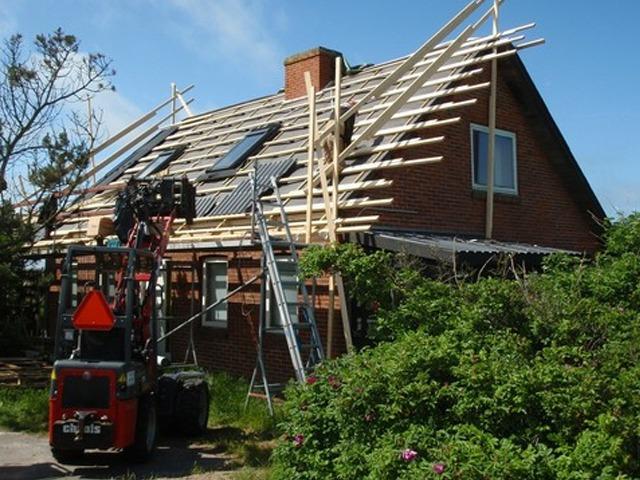 Hus med tagrenovering, stillads og byggeudstyr, omkranset af grønne planter og træer.
