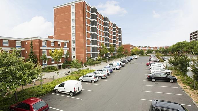 En parkeringsplads med biler foran en moderne bygning med røde mursten og grønne træer i baggrunden.