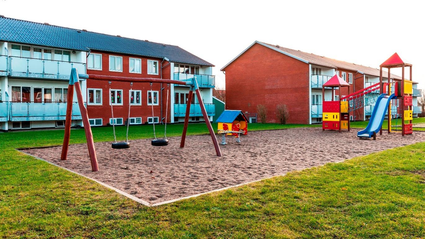 Legestue med gynger, rutsjebane og sandkasse mellem to bygninger i et boligområde.
